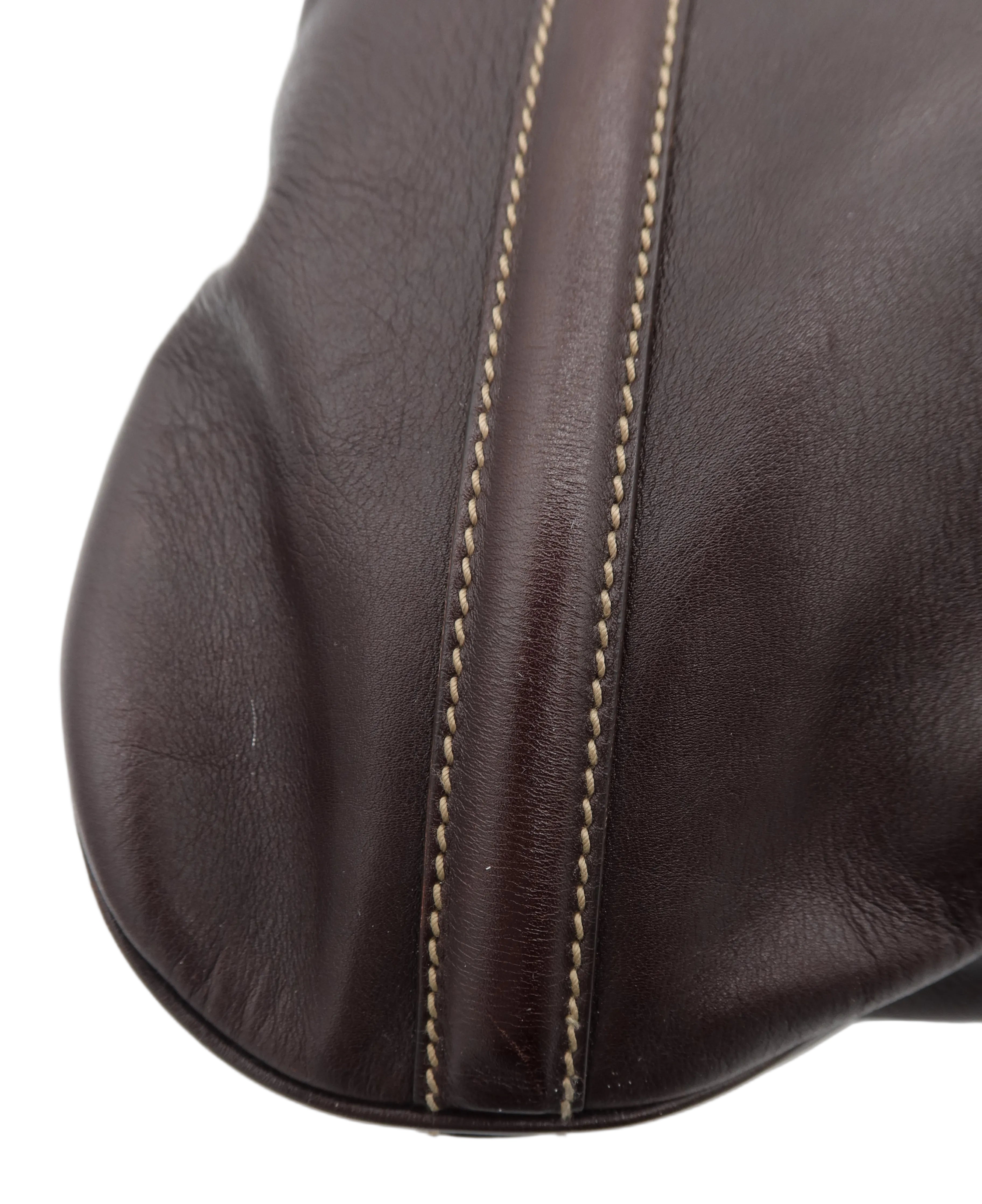 Close-up of a dark brown leather item, likely a handbag or leather accessory, showing the texture of the leather and a contrasting light beige stitching detail along a seam, set against a plain white background.