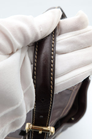 Close-up of a dark brown leather handbag strap with gold-tone hardware, held by a hand wearing a white fabric glove against a light gray background.