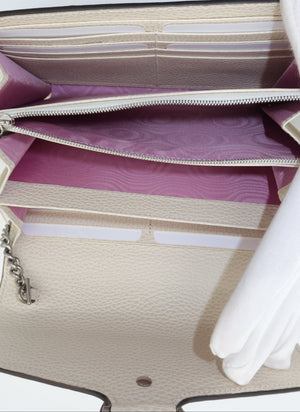 Close-up of a beige pebbled leather clutch or wallet with a silver zipper, featuring a purple patterned interior lining and white fabric gloves holding it open.
