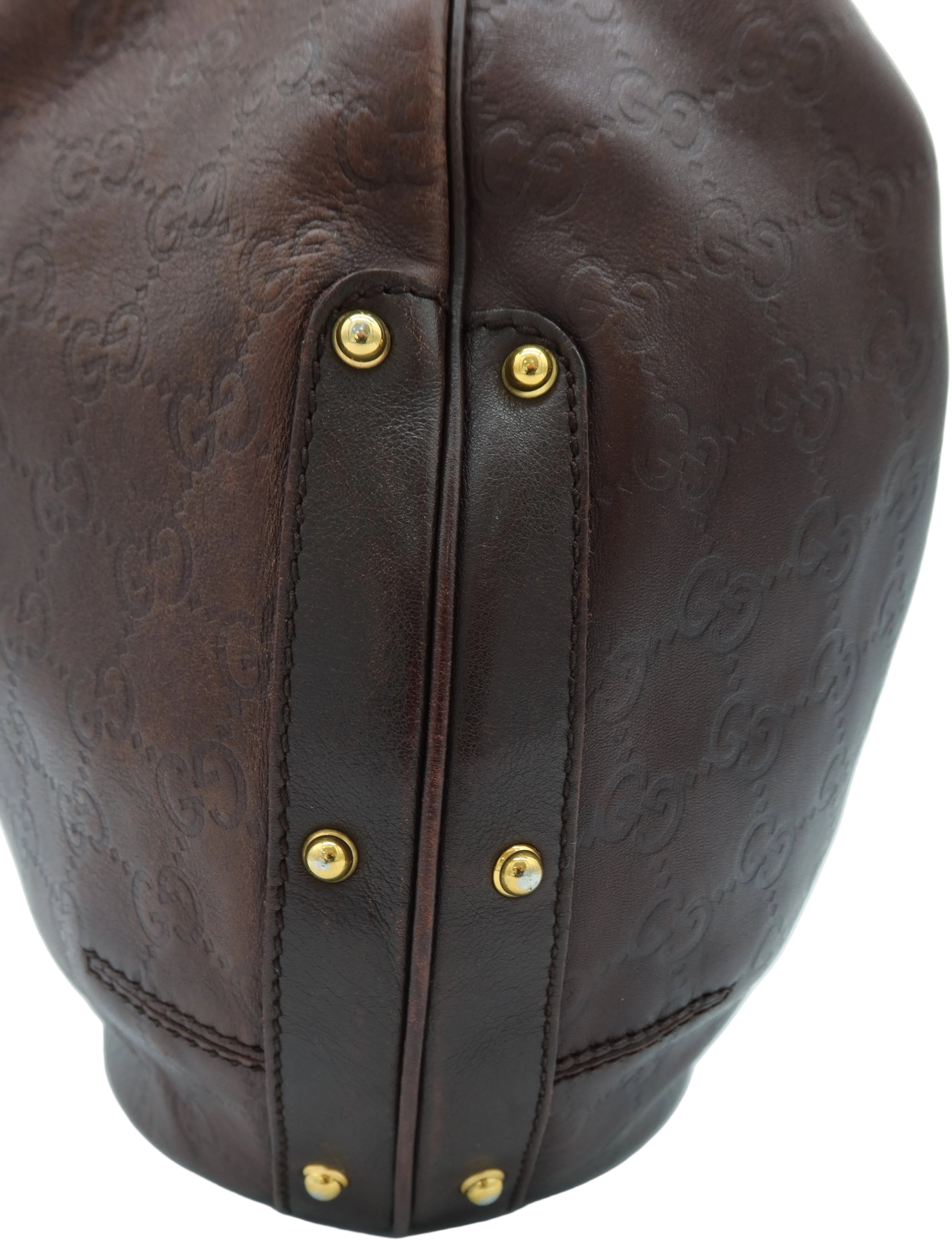 Close-up of a dark brown leather handbag featuring an embossed monogram pattern and gold-tone metal rivets on a light gray background.