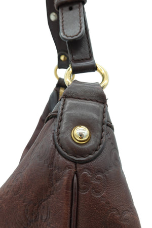Close-up of a dark brown embossed leather handbag showing a gold-tone snap button closure and a matching leather strap with gold hardware against a light background.