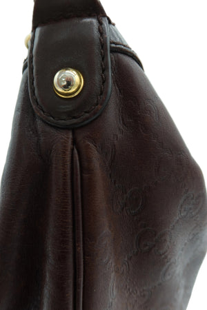 Close-up of a dark brown leather shoulder bag with an embossed monogram pattern and a gold-tone snap button closure.