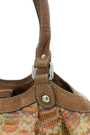 Close-up detail of a handbag showing a brown leather strap with silver-tone hardware and a multicolored woven fabric body against a light gray background.