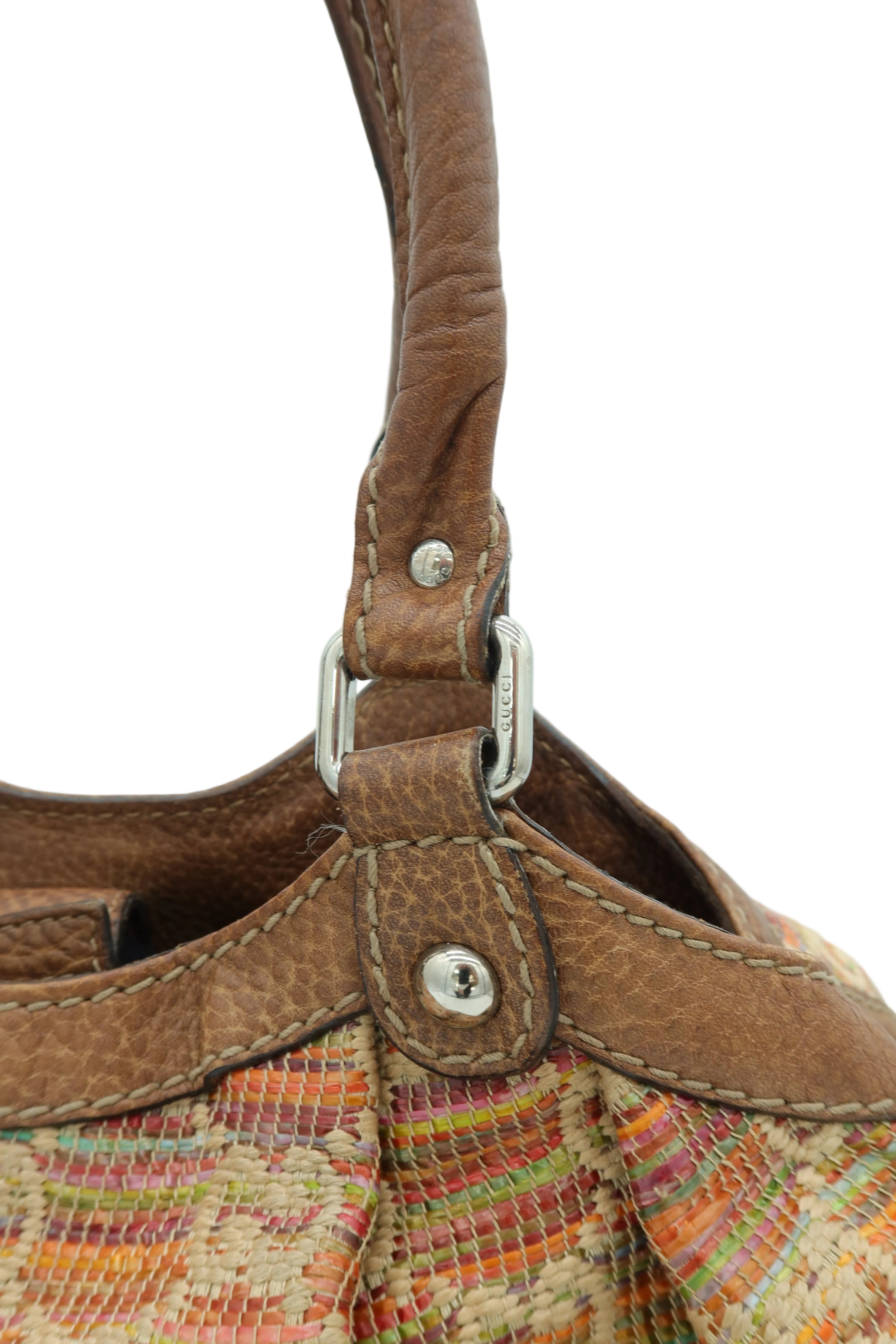 Close-up detail of a handbag showing a brown leather strap with silver-tone hardware and a multicolored woven fabric body against a light gray background.