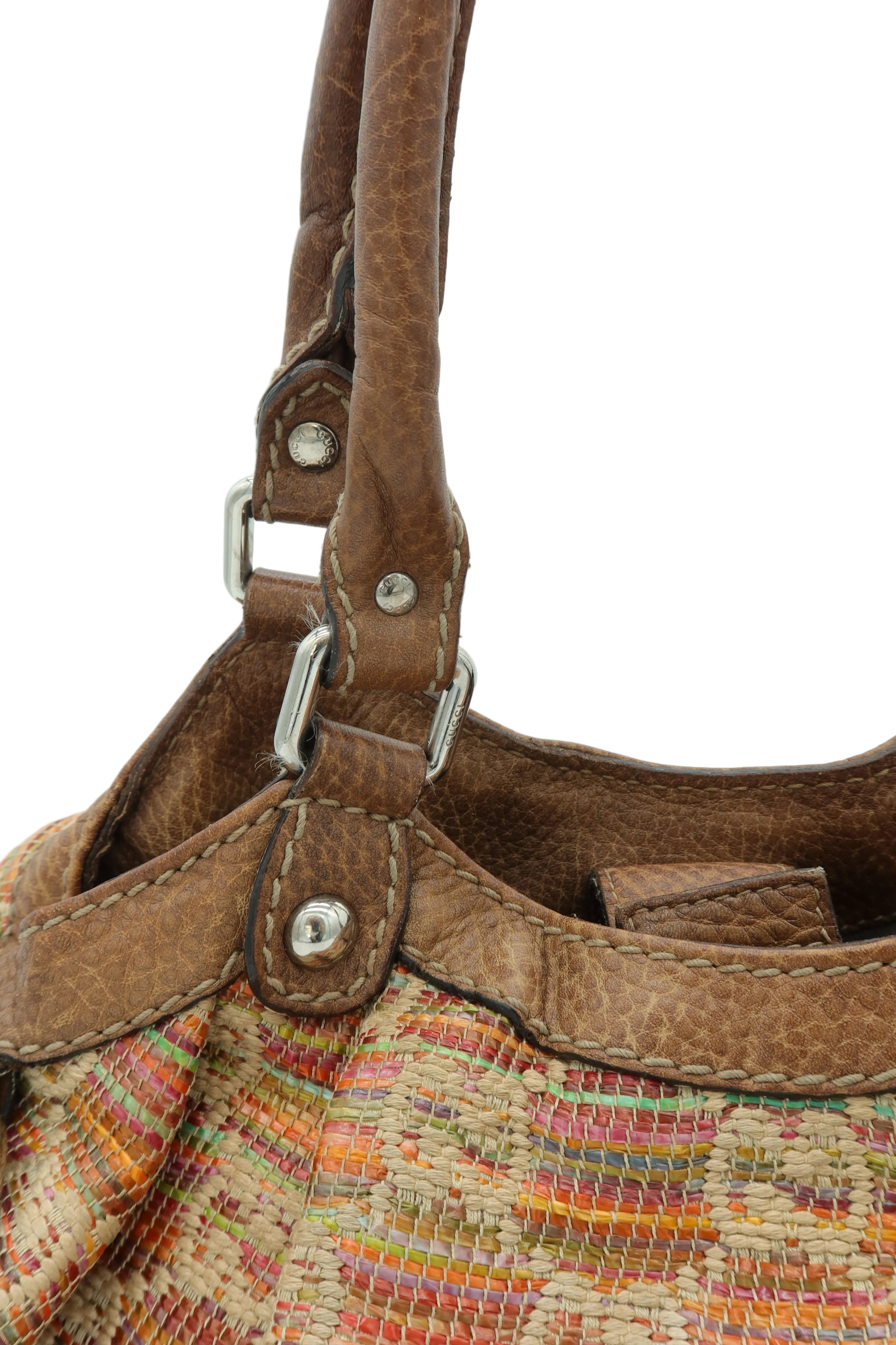 Close-up of a handbag showing brown leather handles and trim with silver-tone hardware, attached to a woven multicolor body in beige, orange, pink, and green tones against a light gray background.
