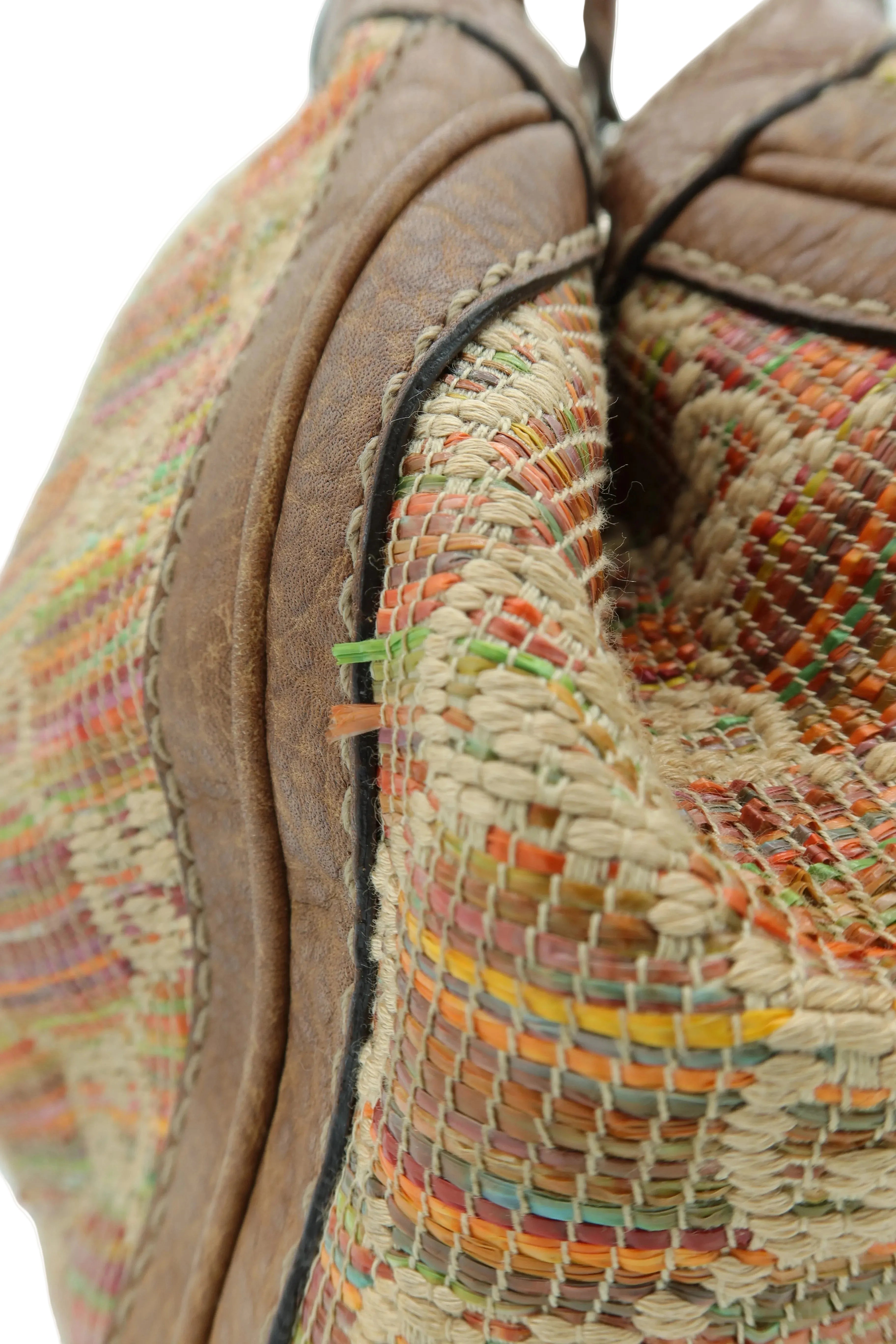 Close-up detail of a woven straw and leather bag corner, showing a beige and brown textured body with multicolored woven stripes and black trim.