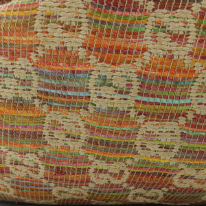 A close-up of a woven textile or rug showing a multicolor pattern of orange, red, beige, yellow, green, and blue threads, resting on a white surface.