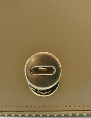 Close-up of a gold-tone metal turn-lock clasp on a tan leather surface, with a small Ferragamo script logo visible on the clasp base.