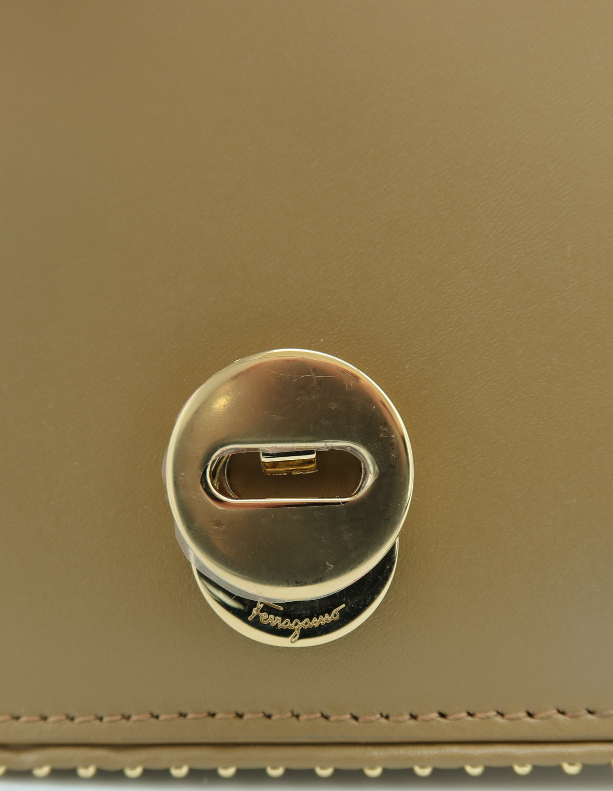 Close-up of a gold-tone metal turn-lock clasp on a tan leather surface, with a small Ferragamo script logo visible on the clasp base.
