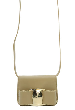 A small structured shoulder bag in light beige leather with a prominent gold-tone bow detail and strap on a light blue background.