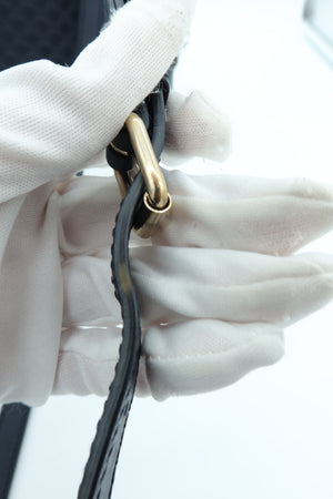A close-up of a hand wearing white gloves holding a black leather strap with a polished gold-tone metal ring and D-ring hardware against a bright white background.