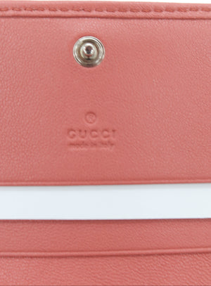 Close-up of a pink leather wallet or clutch with a white horizontal stripe and a silver snap button, featuring an embossed Gucci logo.
