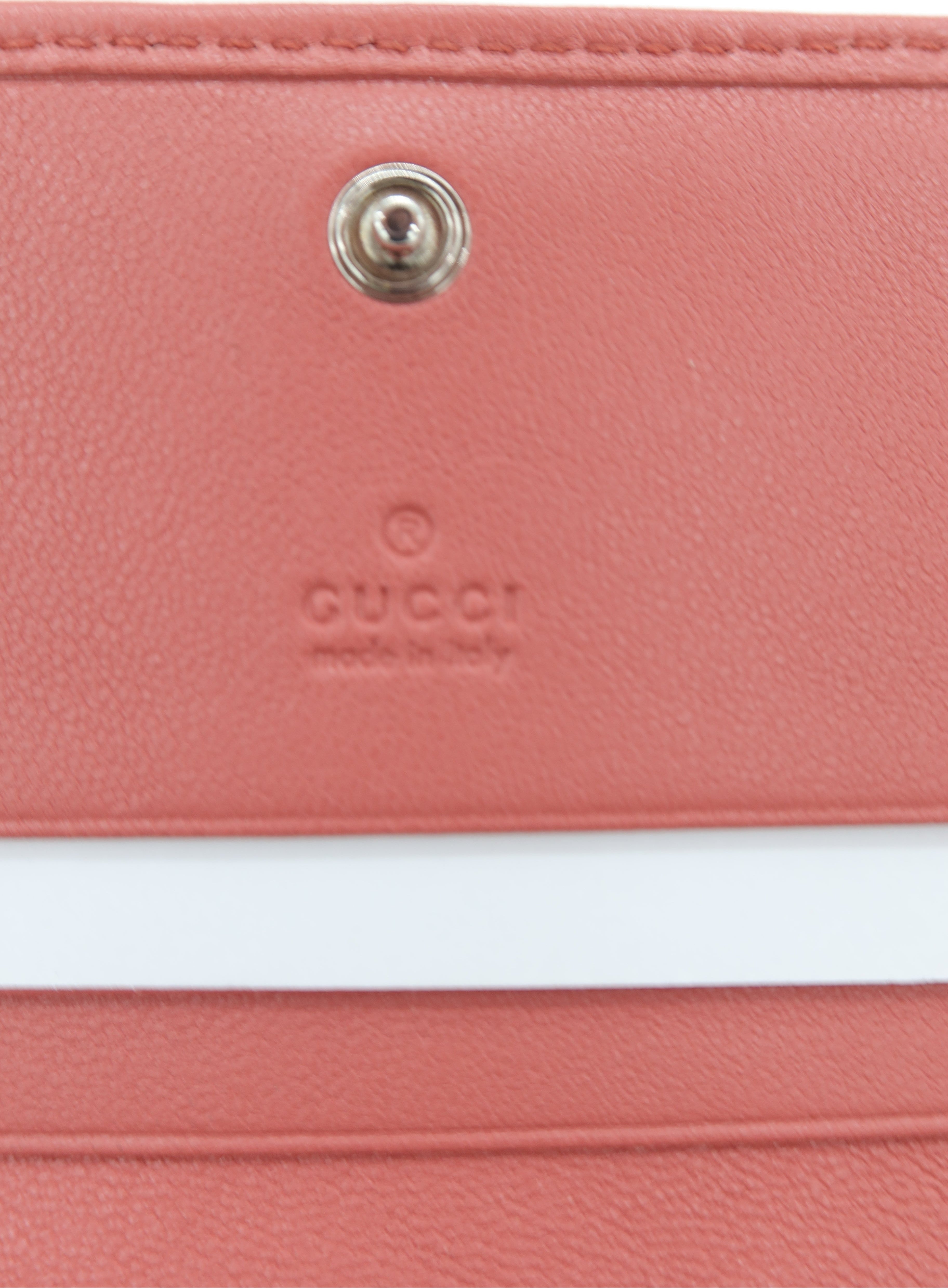 Close-up of a pink leather wallet or clutch with a white horizontal stripe and a silver snap button, featuring an embossed Gucci logo.