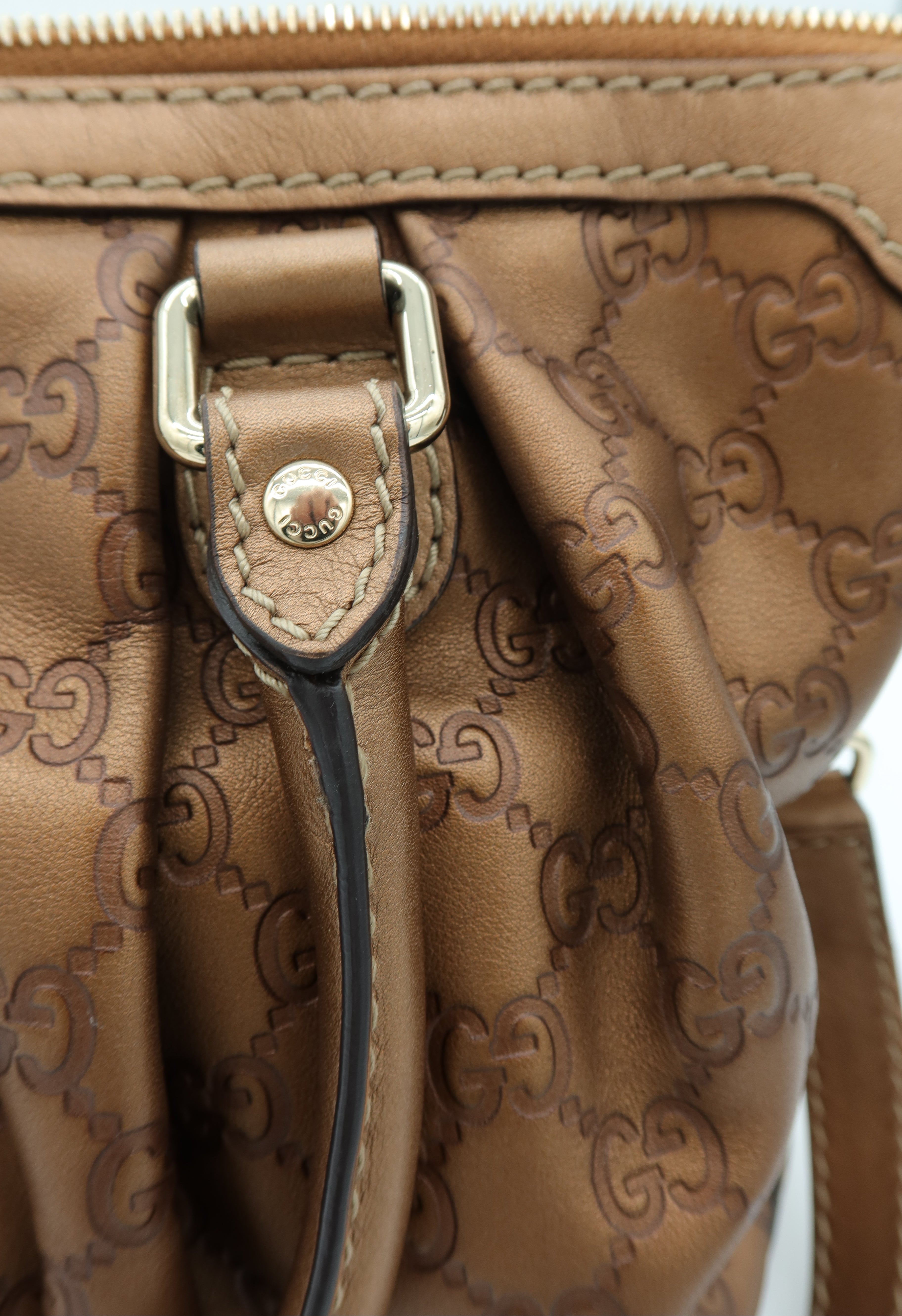 Close-up of a brown leather handbag with an embossed monogram pattern, a gold-tone metal buckle, and a black leather strap detail.