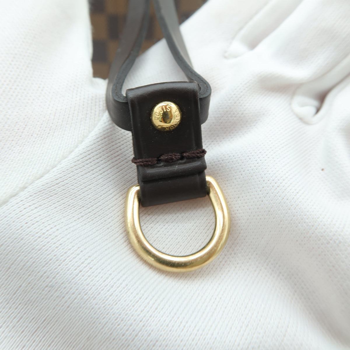 Close-up of a dark brown leather strap with a gold-tone D-ring and a small gold snap button, resting on a white textured surface with a brown checkered pattern visible in the background.