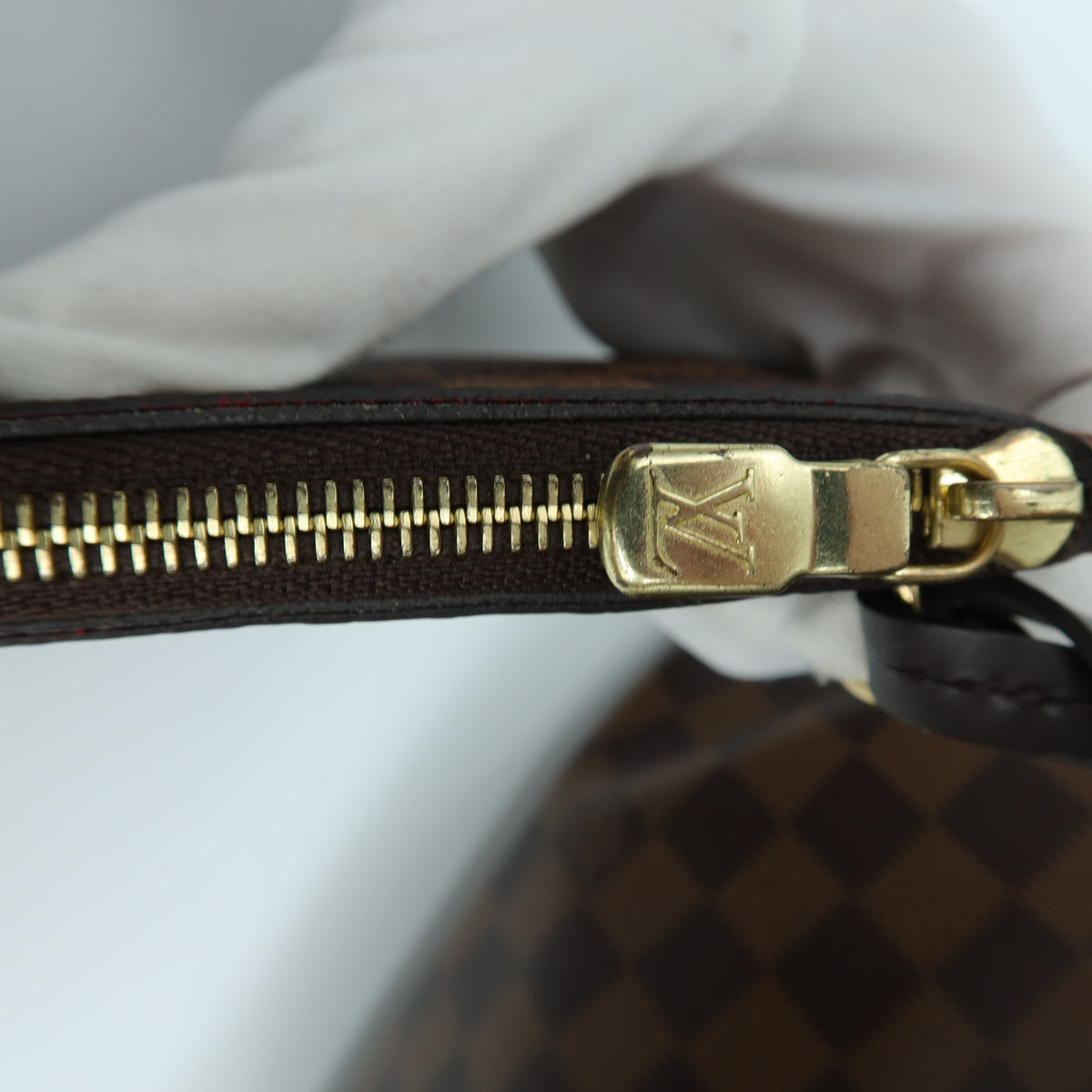 Close-up of a Louis Vuitton Damier canvas bag with a brown and black checkered pattern, featuring a gold-tone metal zipper with a gold LV logo pull, held by a white gloved hand.
