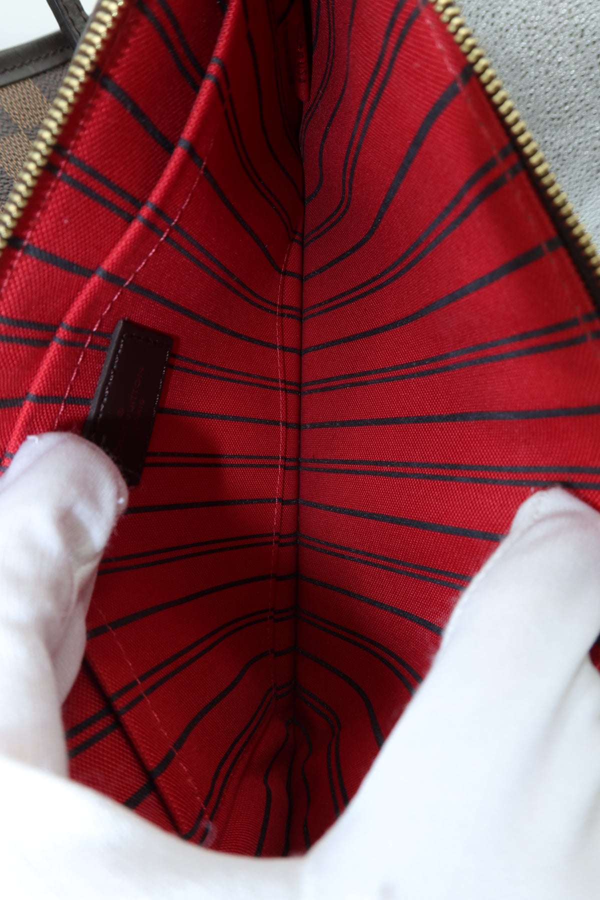 Close-up of the interior of a red fabric bag with black plaid stripes, held open by white-gloved hands. A gold zipper runs along the top edge, and a small dark brown leather brand tab is visible on the left side.