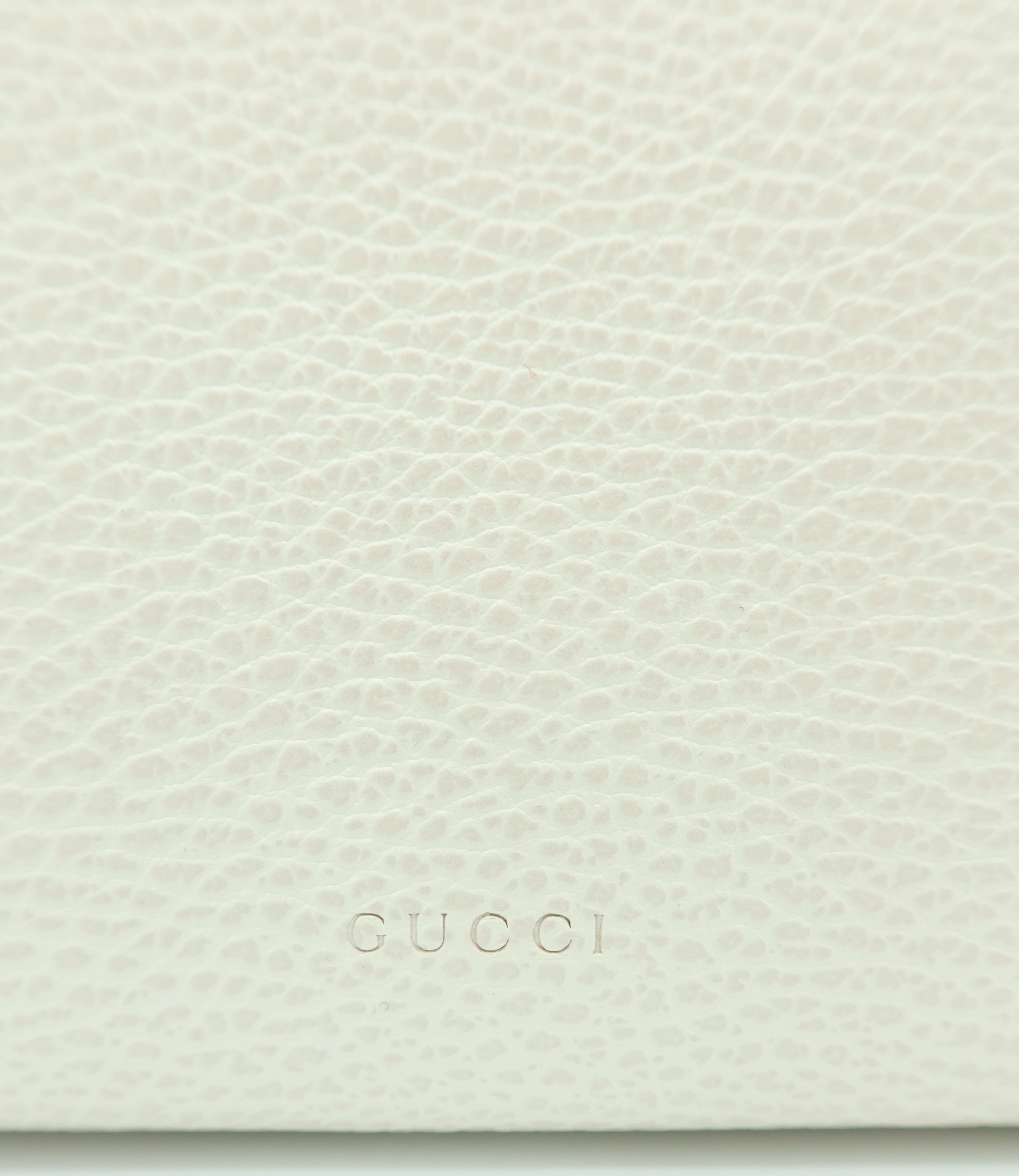 Close-up of a light cream pebbled leather surface with the embossed brand name GUCCI centered near the bottom.