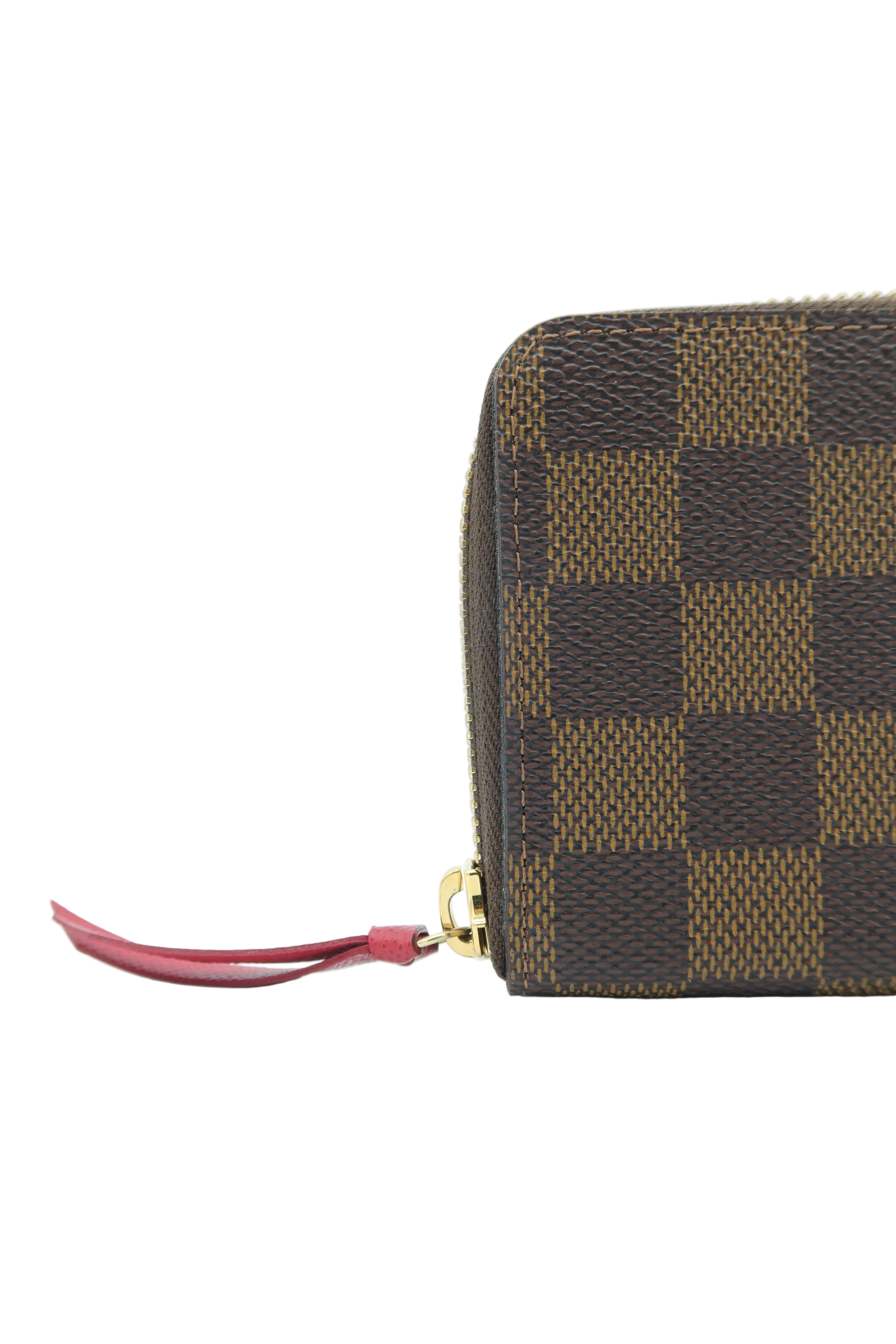 A close-up of a Louis Vuitton zip-around wallet in Damier Ebene canvas, showing the brown and tan checkered pattern and a red leather zipper pull with a gold-tone metal tip, set against a plain white background.