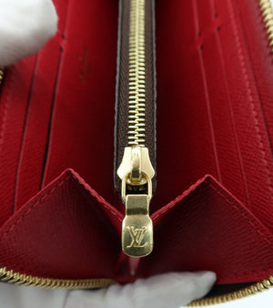 Close-up of a red textured leather wallet or card holder with a gold-tone zipper and an embossed Louis Vuitton monogram on the zipper pull, held by a white-gloved hand.