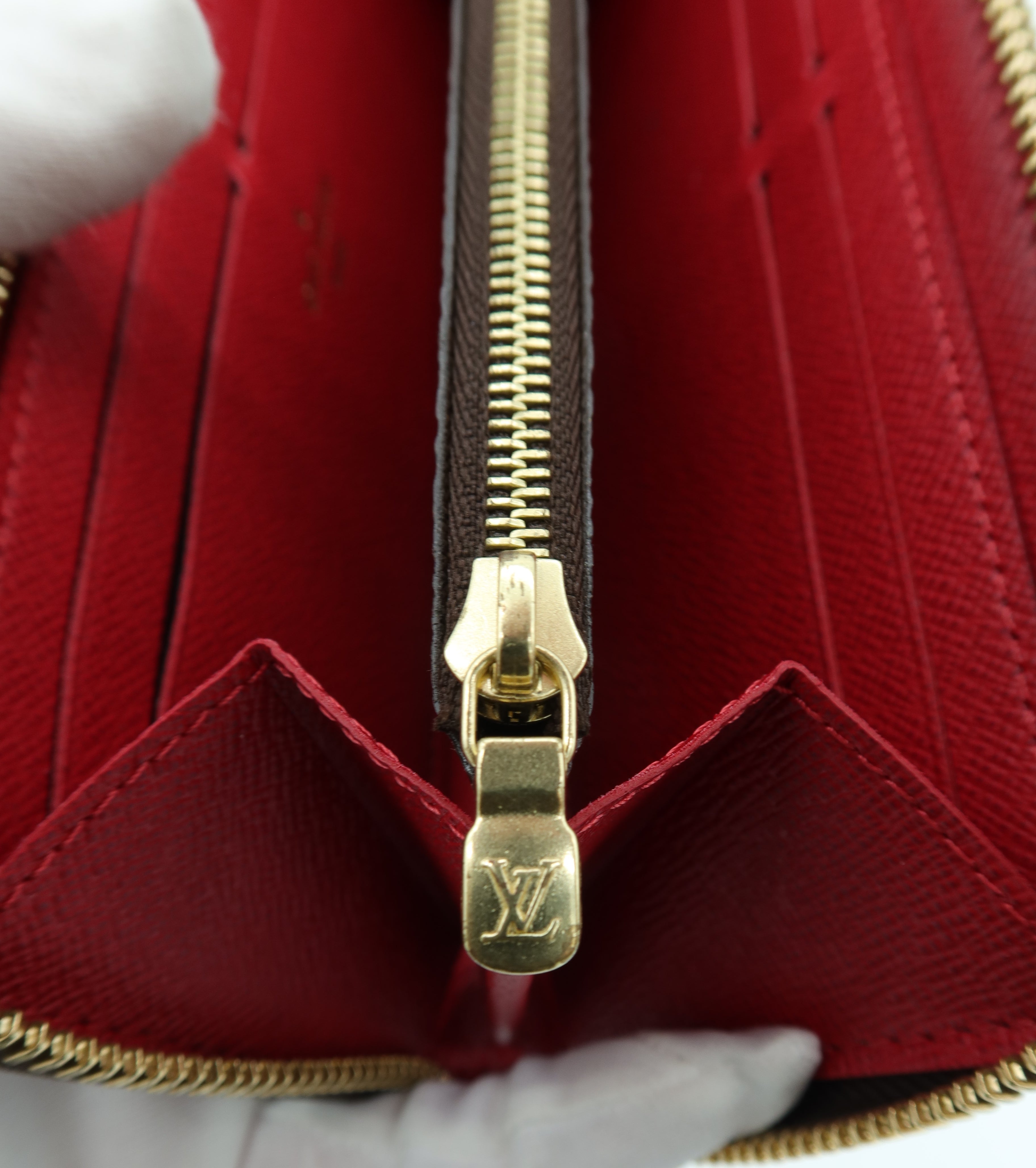 Close-up of a red textured leather wallet or card holder with a gold-tone zipper and an embossed Louis Vuitton monogram on the zipper pull, held by a white-gloved hand.