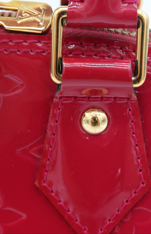 Close-up of a red patent leather handbag with gold-tone hardware, showing a strap, buckle, and a small LV monogram detail against a white background.