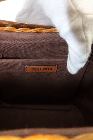 Close-up of the interior of a brown woven bag, showing a small brown leather Miu Miu logo patch and part of a white textured fabric or glove at the top.