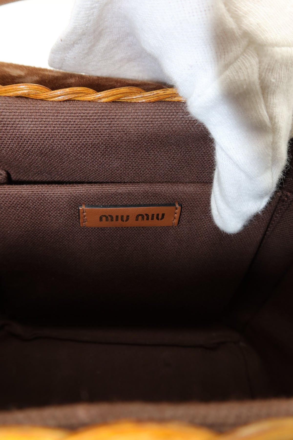 Close-up of the interior of a brown woven bag, showing a small brown leather Miu Miu logo patch and part of a white textured fabric or glove at the top.
