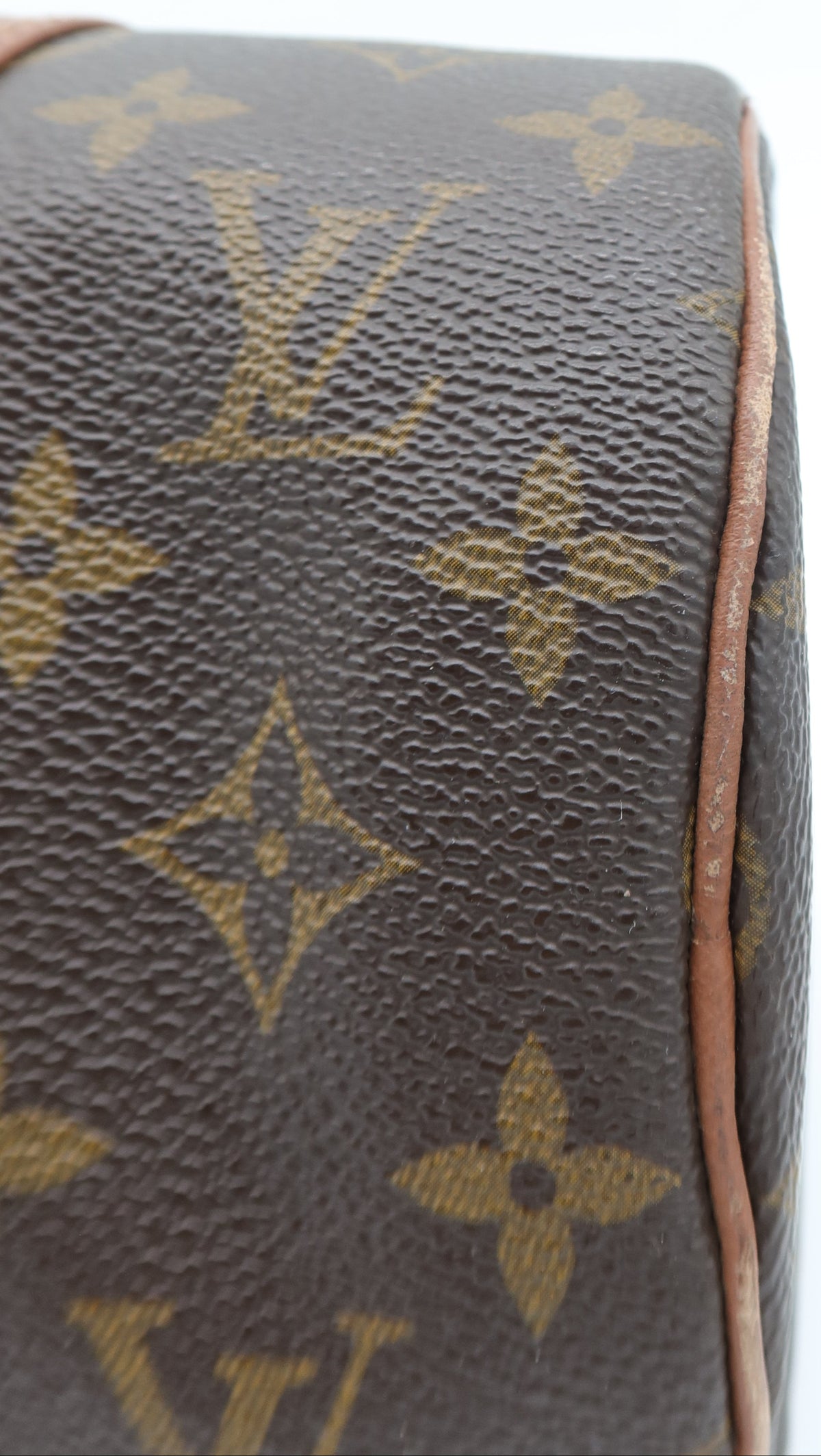 Close-up of a Louis Vuitton monogram canvas bag corner showing the classic brown and tan LV pattern with a light brown leather edge on a white background.