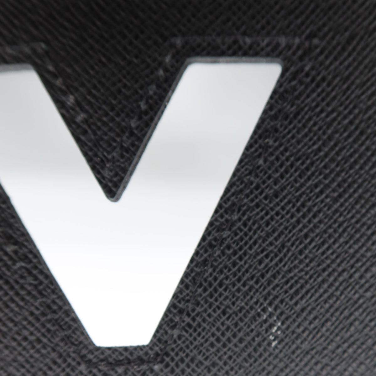 Close-up of a black textured leather item featuring a large, polished silver metallic 'V' logo, with a white circular element visible at the top edge.