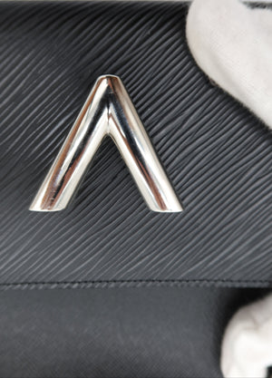 Close-up of a black textured leather clutch or handbag with a prominent polished silver chevron V-shaped metal emblem on the front, with a white gloved hand partially visible holding the item.