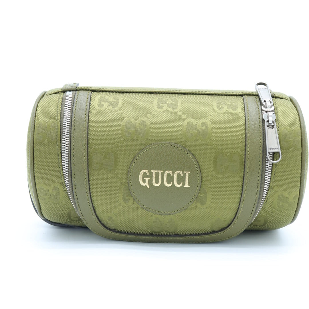 A green Gucci toiletry bag featuring the brand's signature GG monogram pattern, a circular green patch with gold 'GUCCI' lettering, and silver-tone zippers.