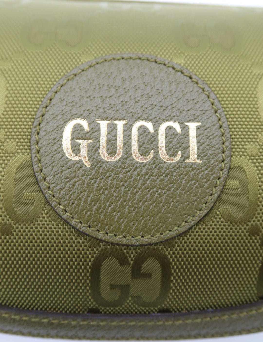 Close-up of a Gucci pouch in olive green GG Supreme canvas with a circular leather patch featuring the brand name 'GUCCI' in gold lettering.