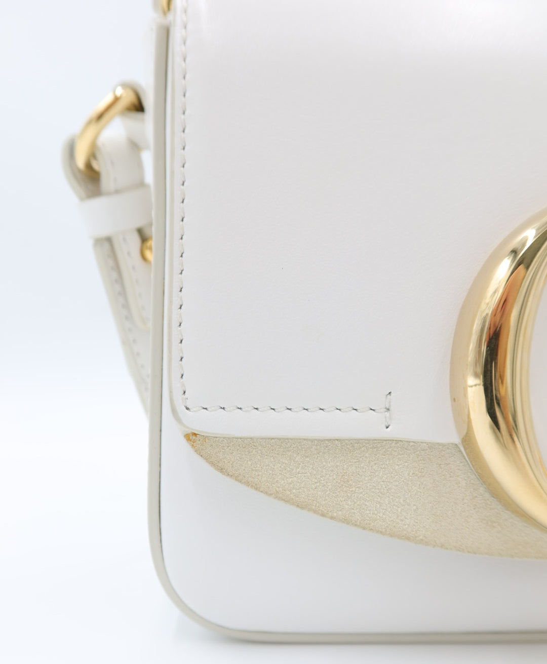 Close-up of a white leather handbag with gold-tone hardware and a light beige suede-like accent panel, photographed on a clean white background.