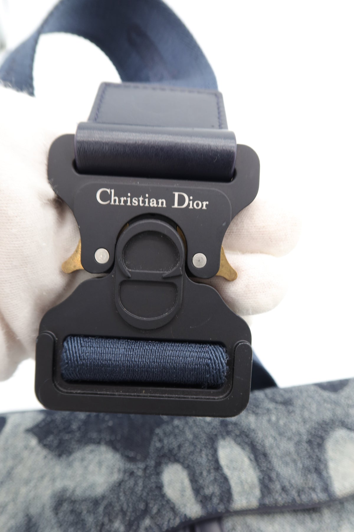 Close-up of a black Christian Dior buckle with a navy blue strap, held by a hand wearing a white glove, with a gray patterned fabric in the background.