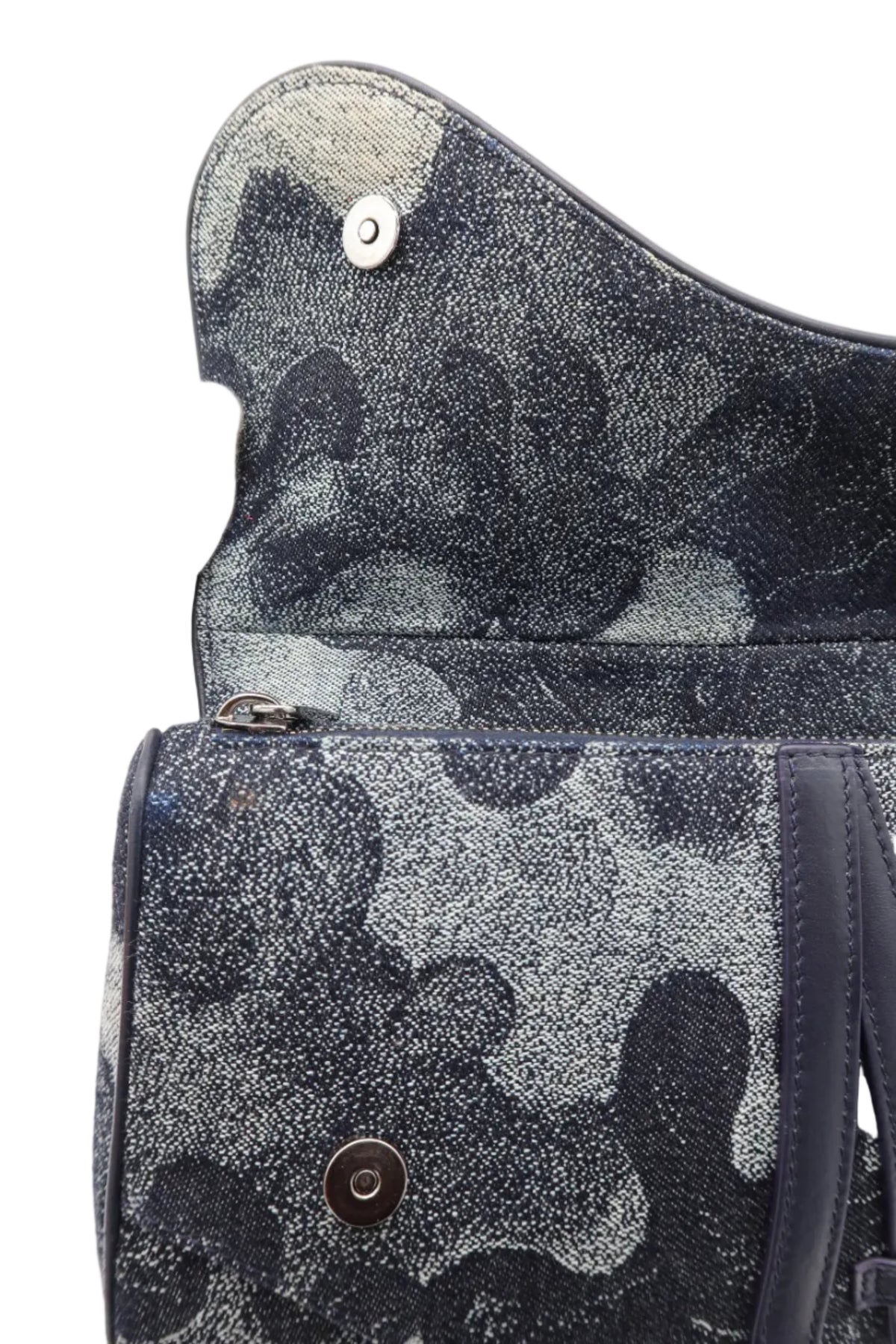 Close-up of a textured handbag interior or flap in a dark abstract camouflage pattern, with a white-gloved hand holding it open against a white background.