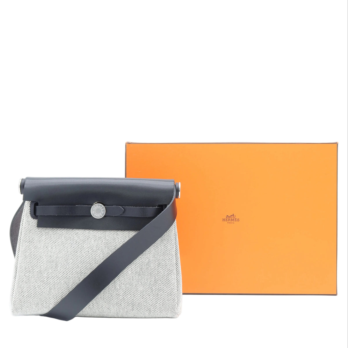 A light gray textured shoulder bag with a black leather flap and strap, shown next to its signature orange Hermès box on a white background.