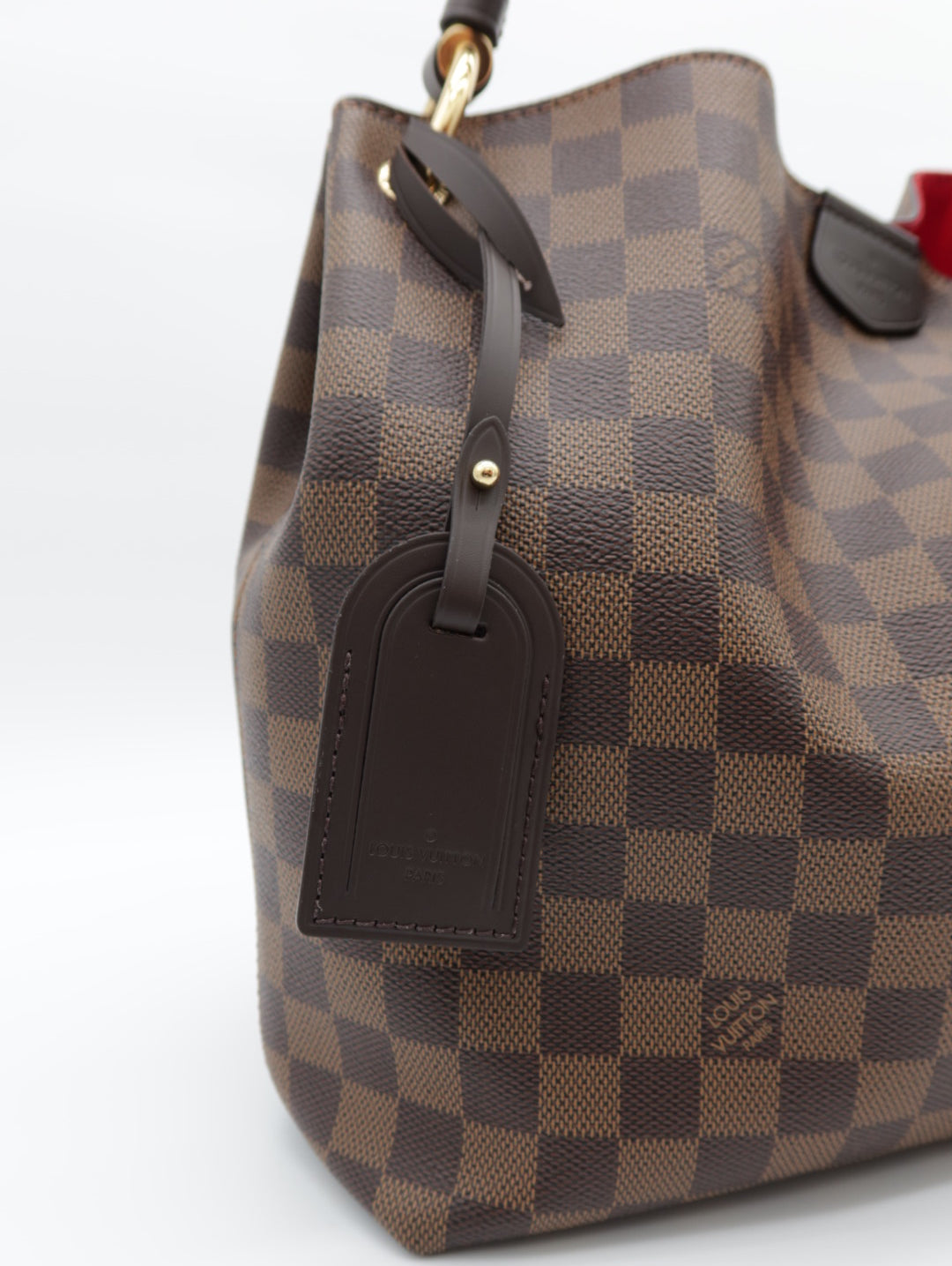 A close-up of a brown and tan checkered canvas handbag with dark brown leather trim and a hanging leather luggage tag, photographed on a white background.