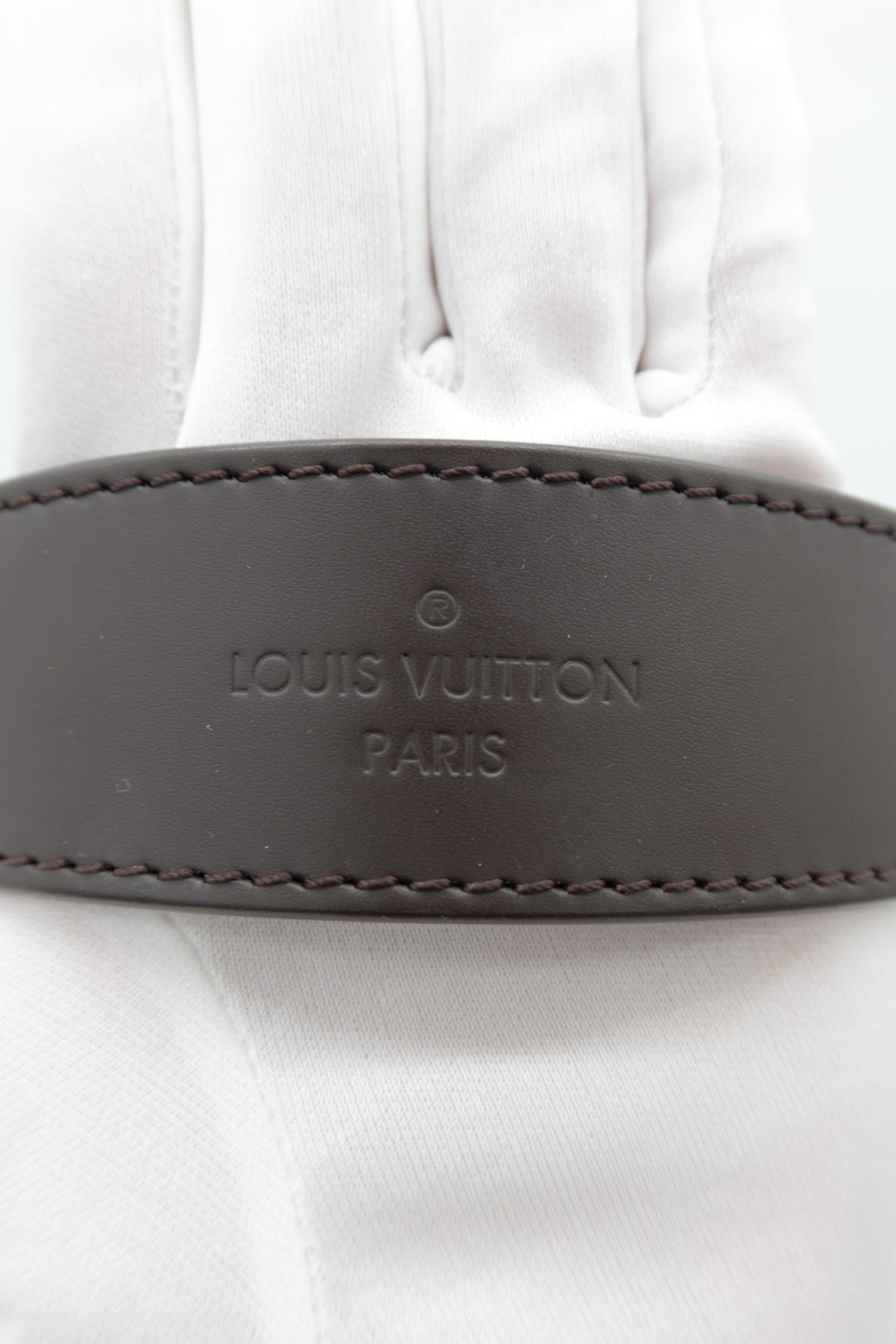 A close-up of a Louis Vuitton leather belt with 'LOUIS VUITTON PARIS' embossed on a white fabric background.