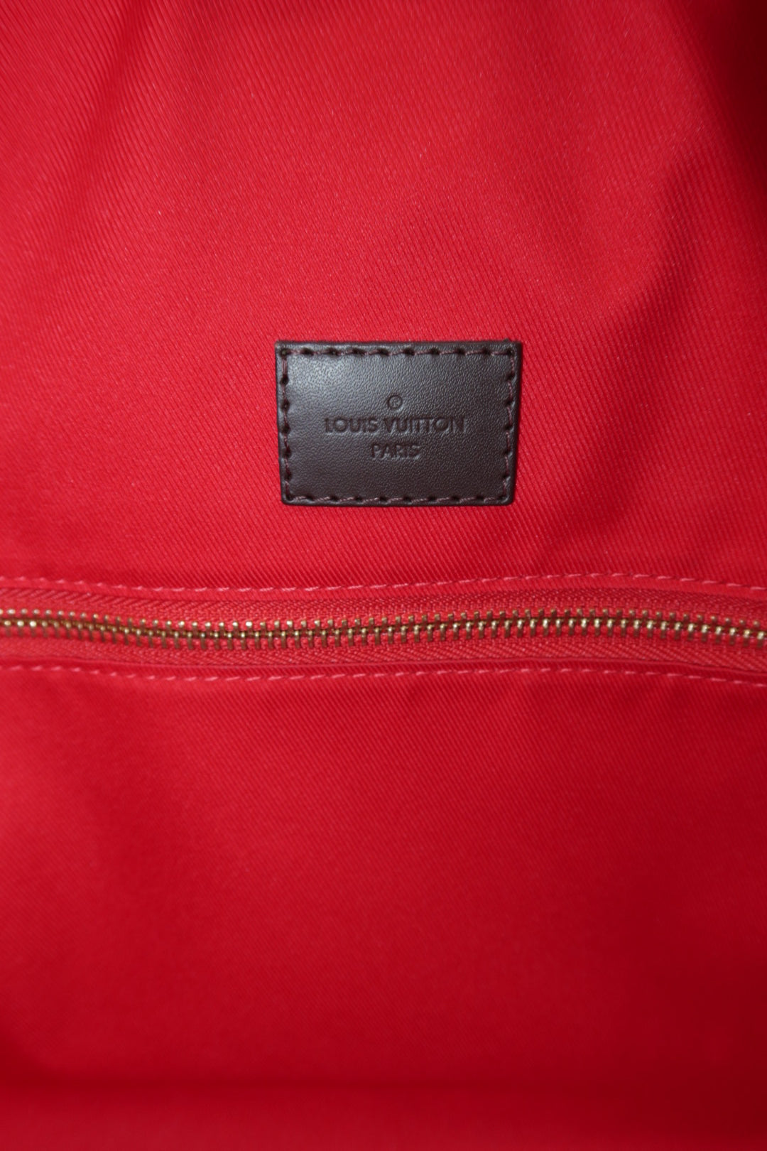 Close-up of a red Louis Vuitton fabric item with a brown leather logo patch reading Louis Vuitton 1947 and a gold zipper.