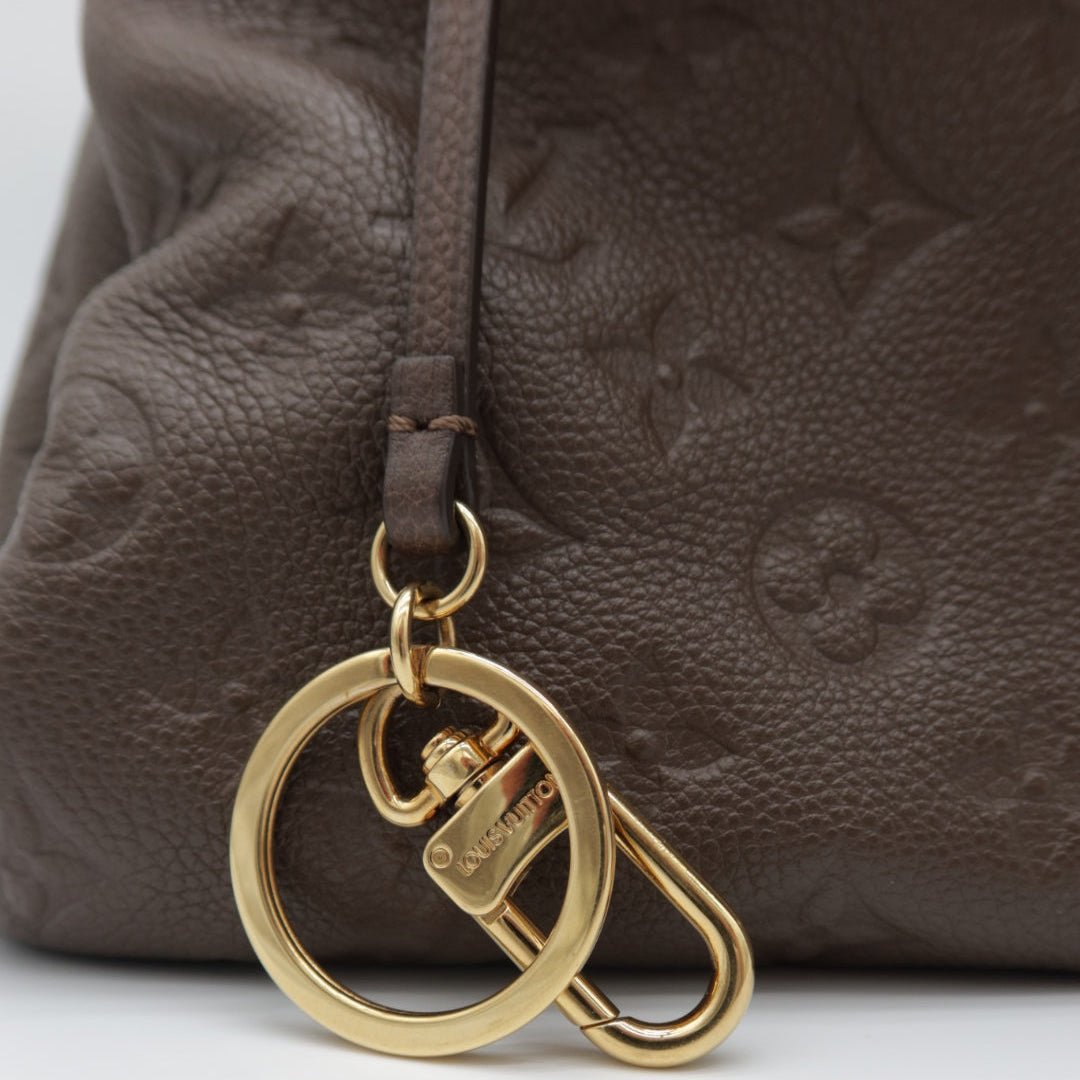 A close-up of a brown textured leather bag featuring an embossed monogram pattern and a gold-tone metal key ring charm with a small lock detail.
