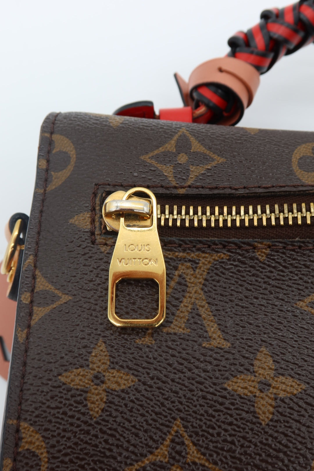 Close-up of a Louis Vuitton monogram canvas bag with a gold-tone zipper pull and a red-and-black braided strap detail on a white background.