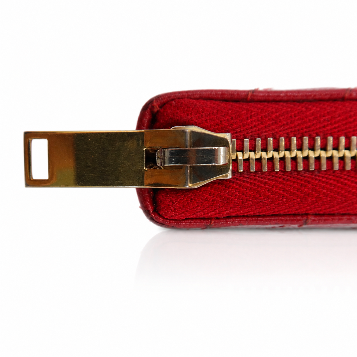 A close-up of a red textured leather zipper pull with a gold-tone metal slider and teeth, set against a plain white background.