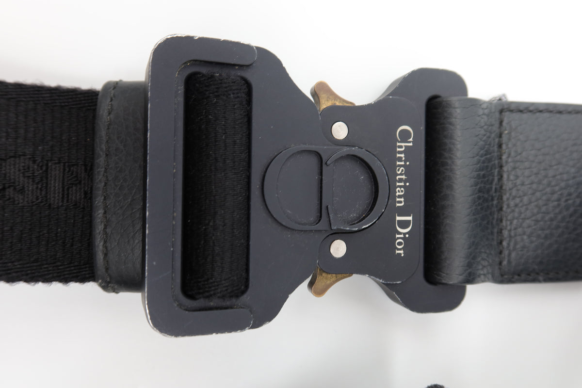 Close-up of a Christian Dior black belt buckle with a black woven strap and textured black leather section, on a white background.