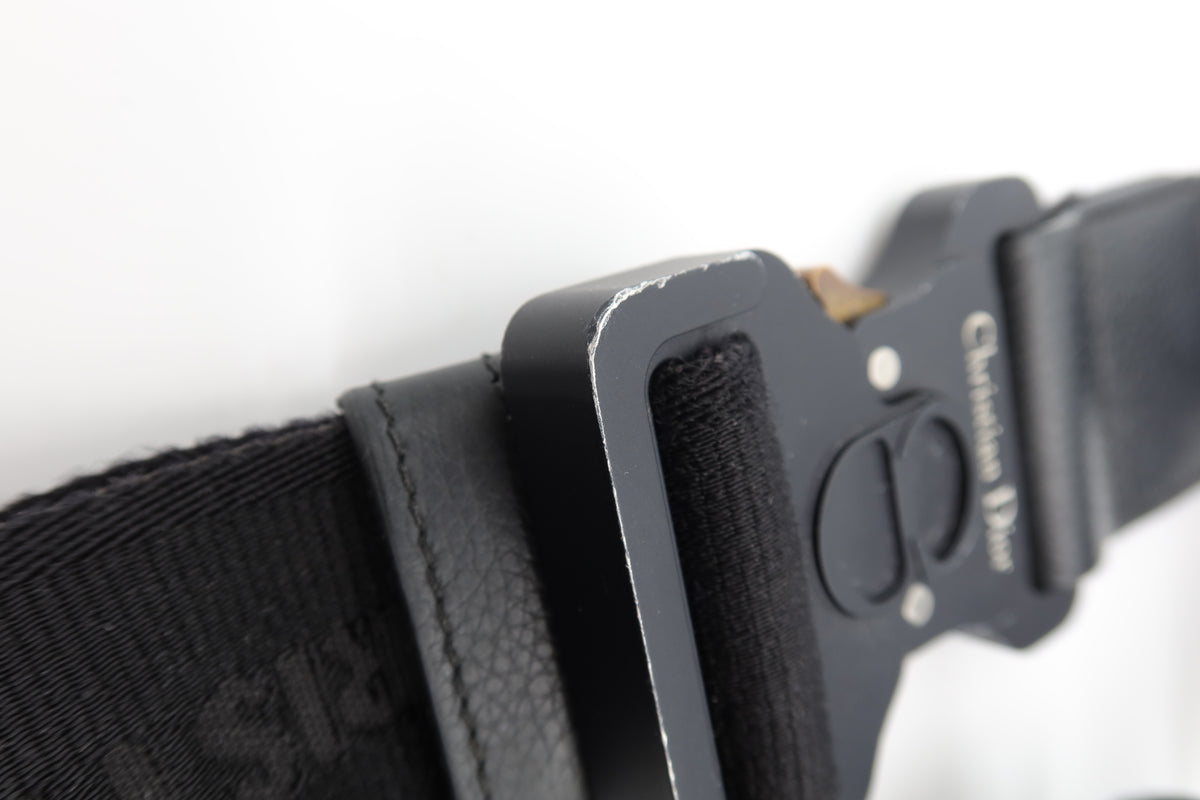 Close-up of a black tactical belt with a matte black metal buckle, featuring a textured webbing strap and a black velcro fastening system. The buckle has 'Christian Dior' printed in white on the side.