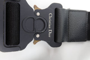 Close-up of a black leather belt buckle with Christian Dior branding and a small gold-tone horsebit detail, on a white background.