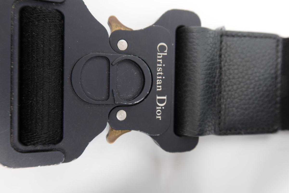 Close-up of a black leather belt buckle with Christian Dior branding and a small gold-tone horsebit detail, on a white background.