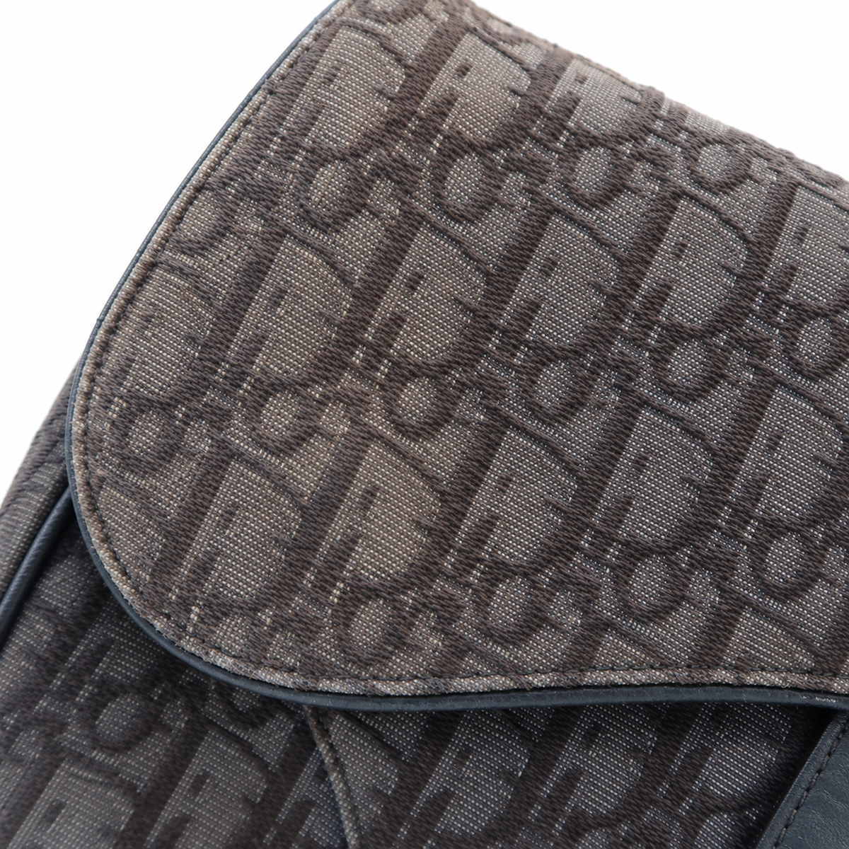 Close-up of a dark brown coated canvas bag featuring the signature Dior Oblique monogram pattern, with a dark leather trim and a white background.