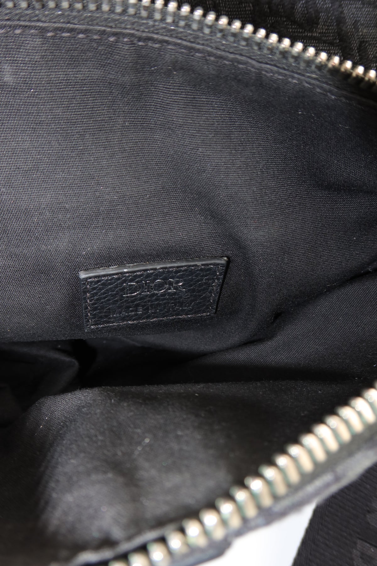 Close-up of the interior of a black Dior bag, showing a black leather brand patch with embossed Dior text and silver-tone zipper trim.