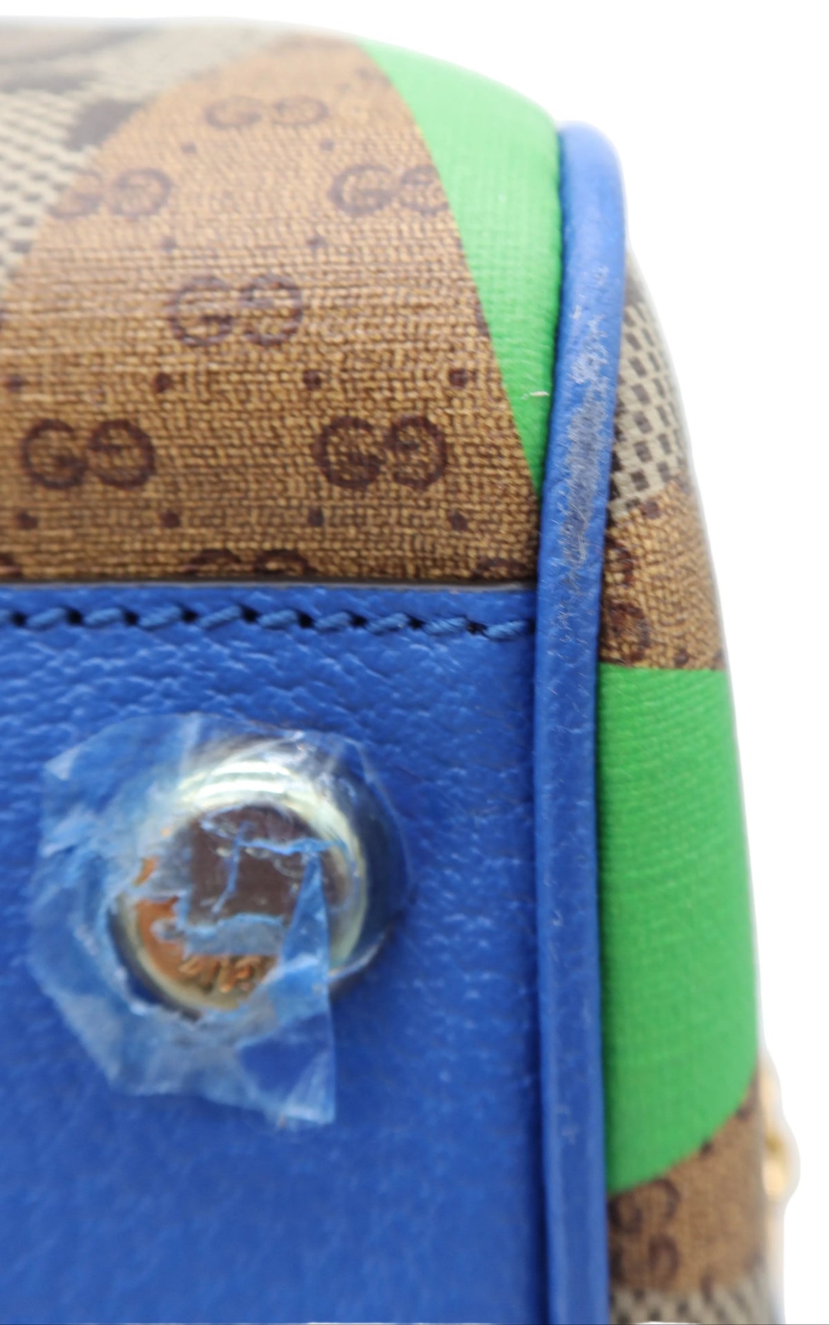 Close-up of a Gucci handbag corner showing brown GG monogram canvas, bright blue leather trim, and green canvas panels on a white background.